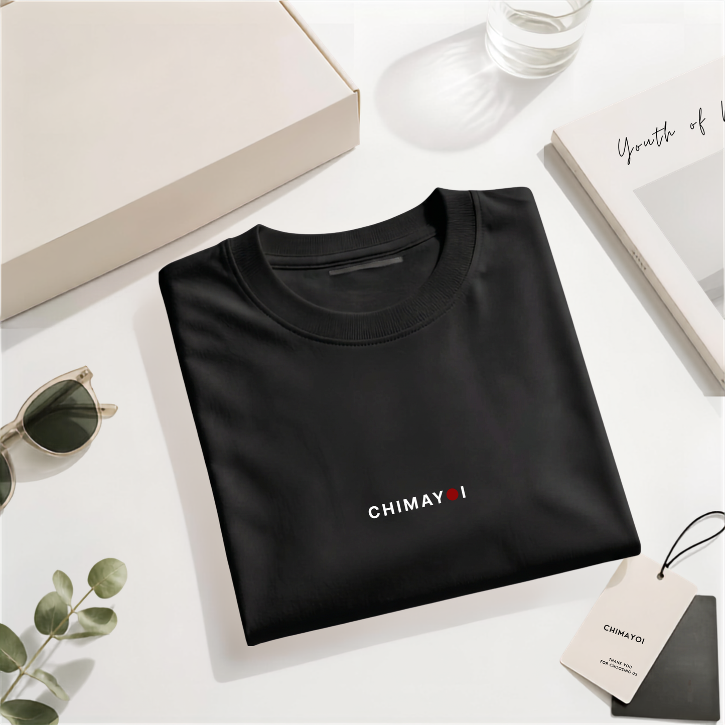 Flat lay of CHIMAYOI Black premium cotton tee folded elegantly with quiet luxury packaging and lifestyle props.