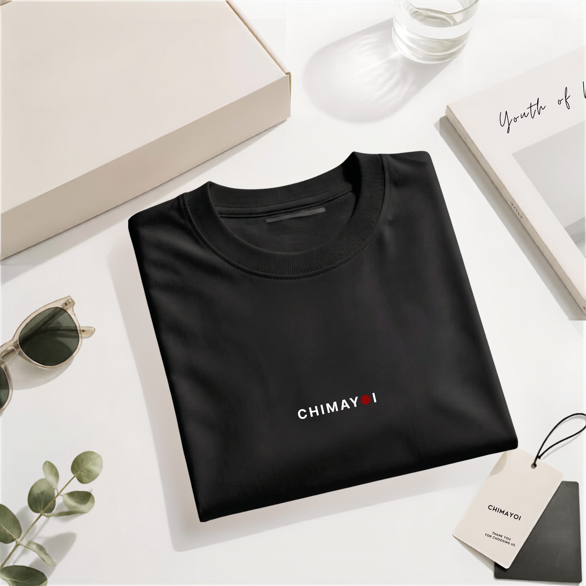 Flat lay of CHIMAYOI Black premium cotton tee folded elegantly with quiet luxury packaging and lifestyle props.